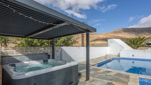 Un jacuzzi y una piscina con pérgola, rodeados por un muro blanco y palmeras.