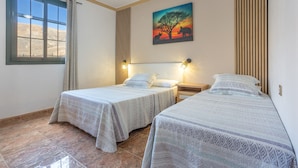 Una habitación con dos camas, una ventana con cortinas, un cuadro de un árbol y animales, y una lámpara fijada a la pared.