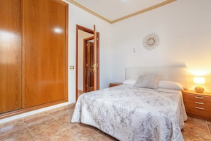Une chambre à coucher avec un lit, une table de chevet, une lampe, une armoire en bois et une porte.