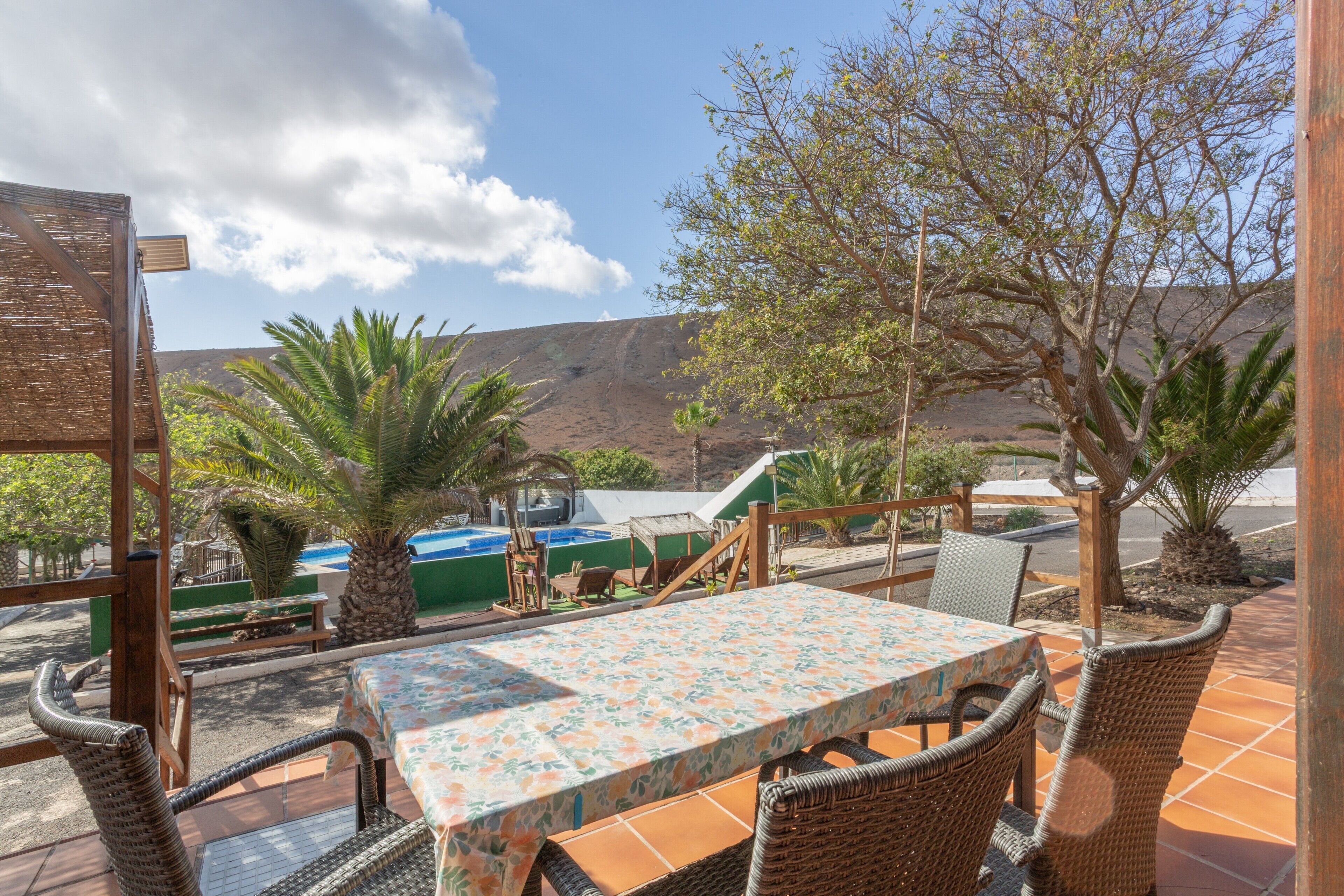 Une terrasse extérieure aménagée avec des meubles en osier, une table recouverte d’une nappe à motifs floraux, et une vue sur une piscine et des montagnes.
