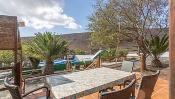 Une terrasse extérieure aménagée avec des meubles en osier, une table recouverte d’une nappe à motifs floraux, et une vue sur une piscine et des montagnes.