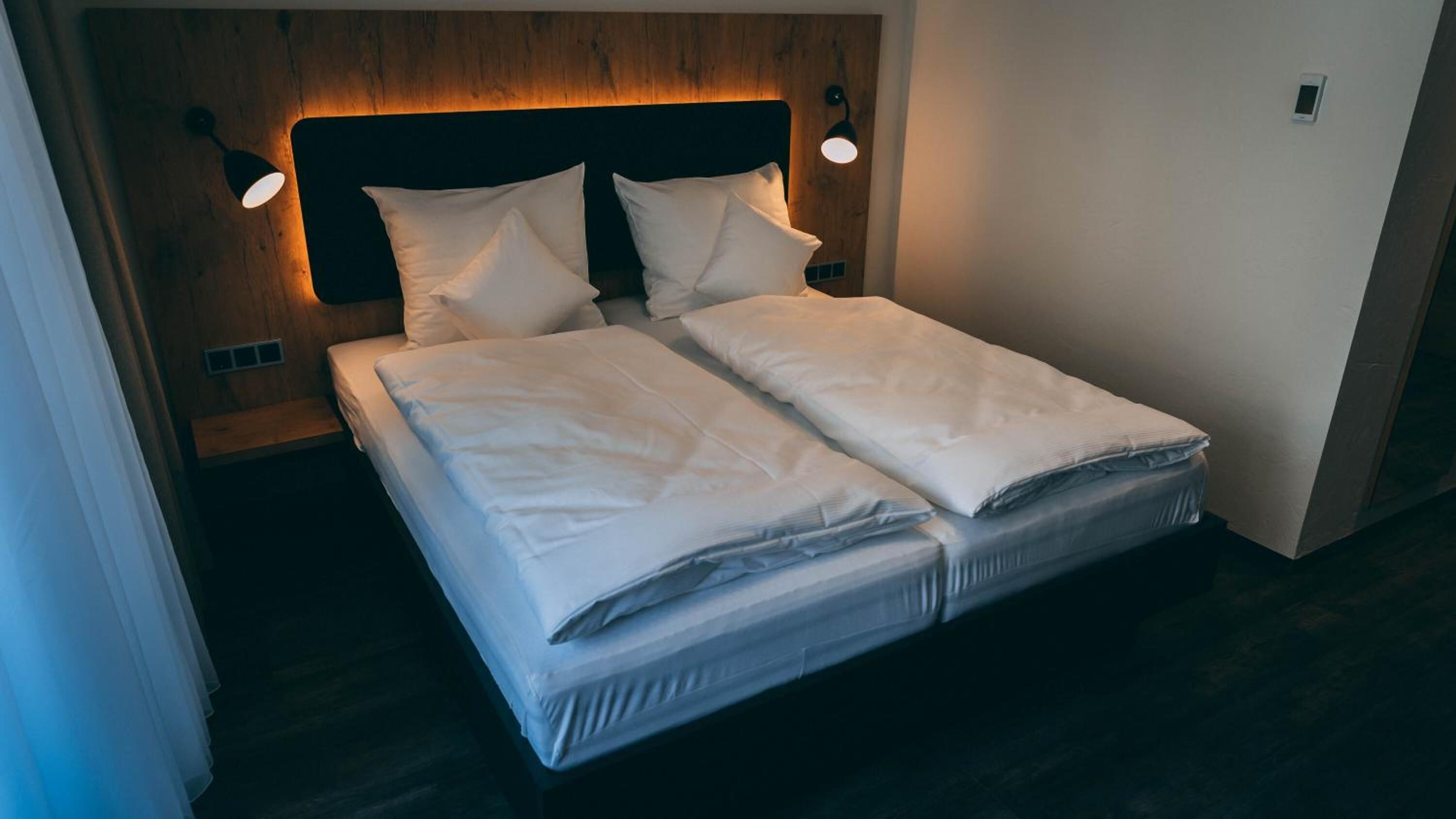 Cama bem arrumada com lençóis brancos em quarto com cabeceira de madeira e duas luminárias de leitura fixadas na parede.