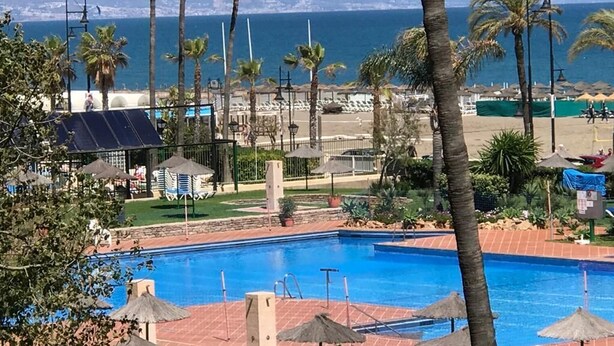 Una piscina con vista al océano, palmeras y una zona de playa con sombrillas.