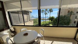 Un balcón con mesa y sillas blancas, vistas a una piscina y palmeras, y un edificio con balcones.
