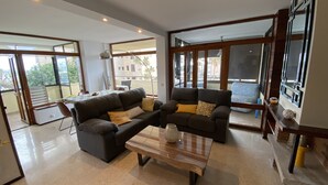 Una sala de estar moderna con un sofá modular, una mesa de centro y un comedor con mesa y sillas. Amplias ventanas ofrecen abundante luz natural y vistas al exterior.