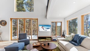 Una sala de estar moderna con chimenea, ventanas amplias y un televisor mostrando a un esquiador.