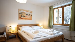 Un letto rifatto con lenzuola bianche e un asciugamano, un comodino con una lampada, un quadro al muro e una finestra con tende.