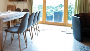 Zona pranzo con tavolo in legno, quattro sedie, televisore a parete e un'ampia finestra con tende.