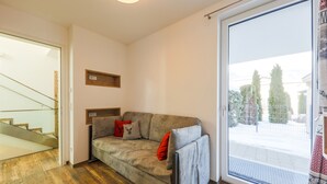 Una sala de estar moderna con un sofá gris, suelo de madera, un ventanal de vidrio que da a un balcón y vistas a árboles y una valla.