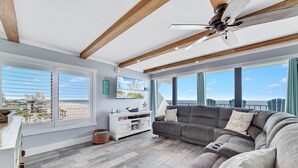 A spacious living room with a gray sectional sofa, a ceiling fan, and large windows offering a view of the beach and cityscape. - Island Winds East 401 | Gulf Front Walk to HangOut | Beach Getaways (Gulf Shores)