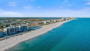 A coastal cityscape with a long sandy beach, a clear blue sea, and a row of high-rise hotels along the shore. - Island Winds East 402 Beachfront Condo | Steps to HangOut! | My Beach Getaways (Gulf Shores)