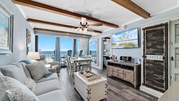 A spacious living area with a high ceiling, wooden beams, a ceiling fan, a dining table with chairs, a coffee station, and a view of the ocean.
