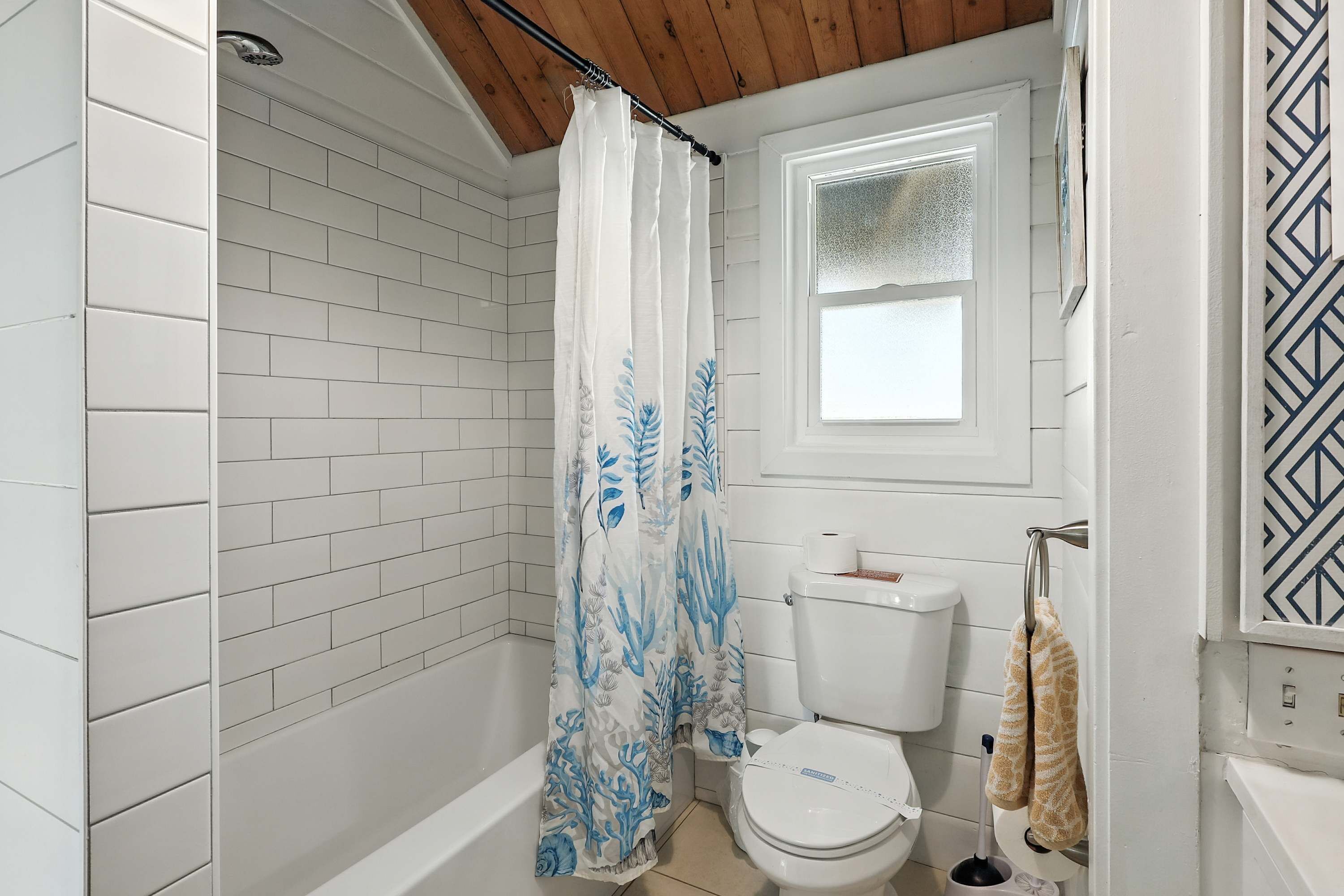 A bathroom with white tiled walls, a shower curtain with blue leaf patterns, a toilet, and a window with frosted glass.