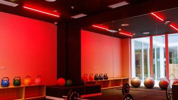 Un gimnasio con pesas de mancuerna y pelotas de ejercicio en estantes, ventanas amplias y techo iluminado con luz roja.