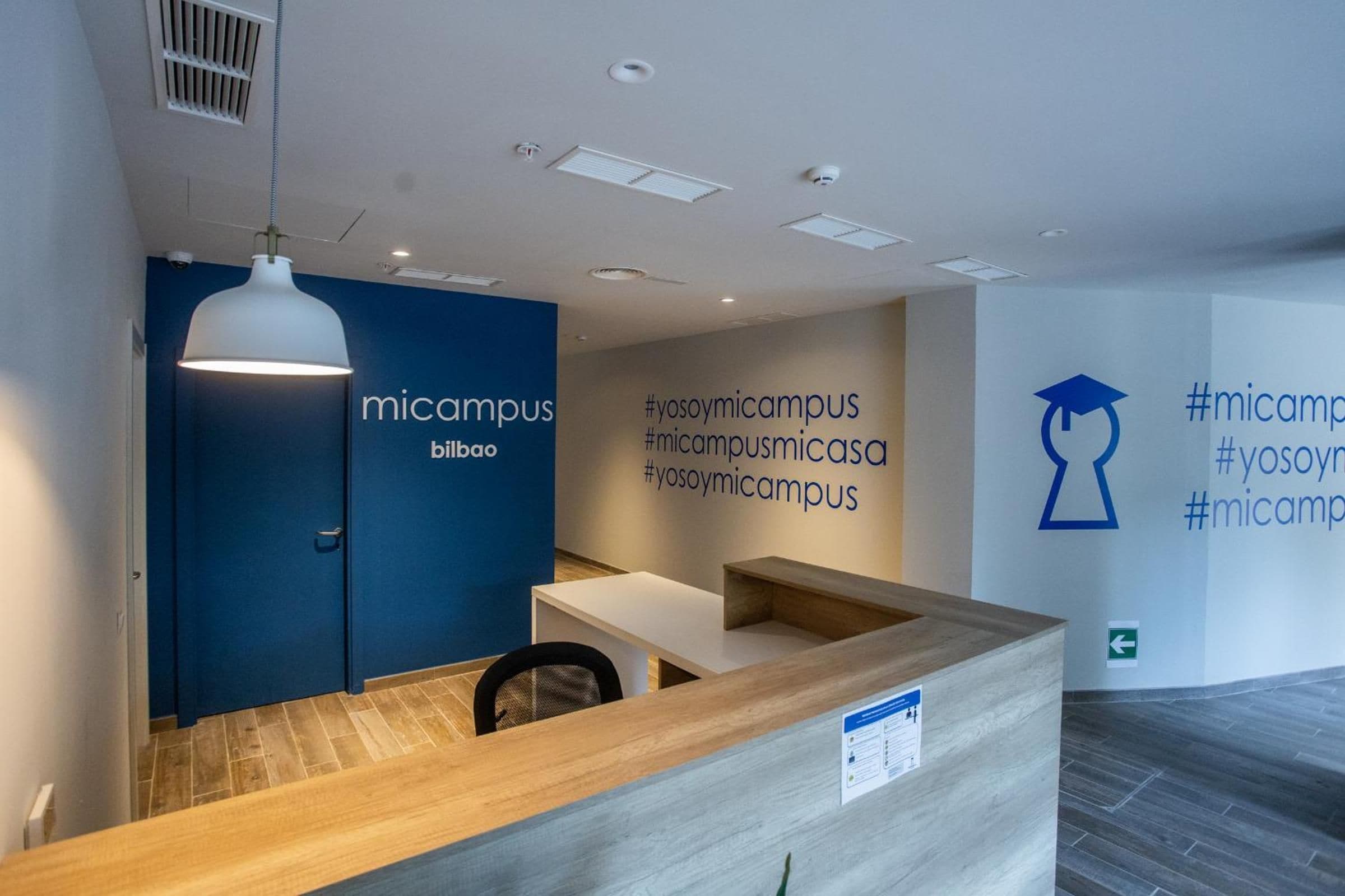 Zona de recepción con pared azul, una lámpara colgante blanca y mostrador de madera.