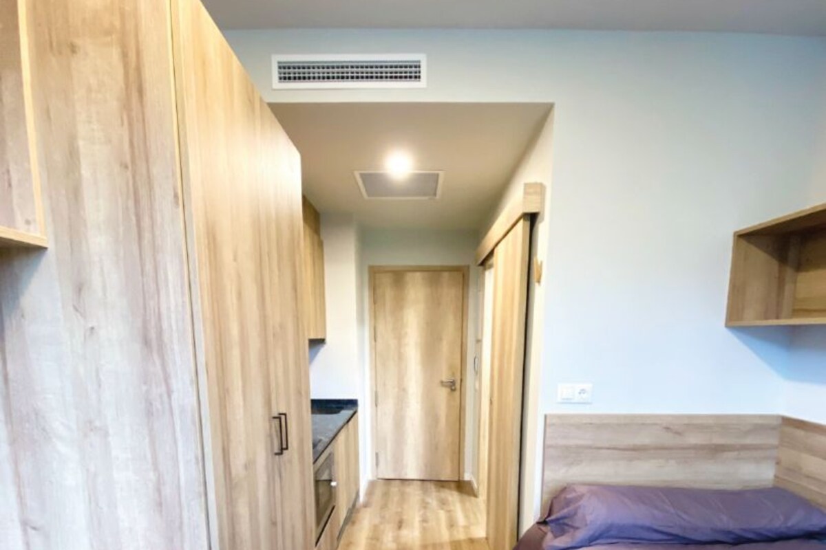 A narrow room with a wooden floor, a bed with purple bedding, a desk with a chair, and a kitchenette area.