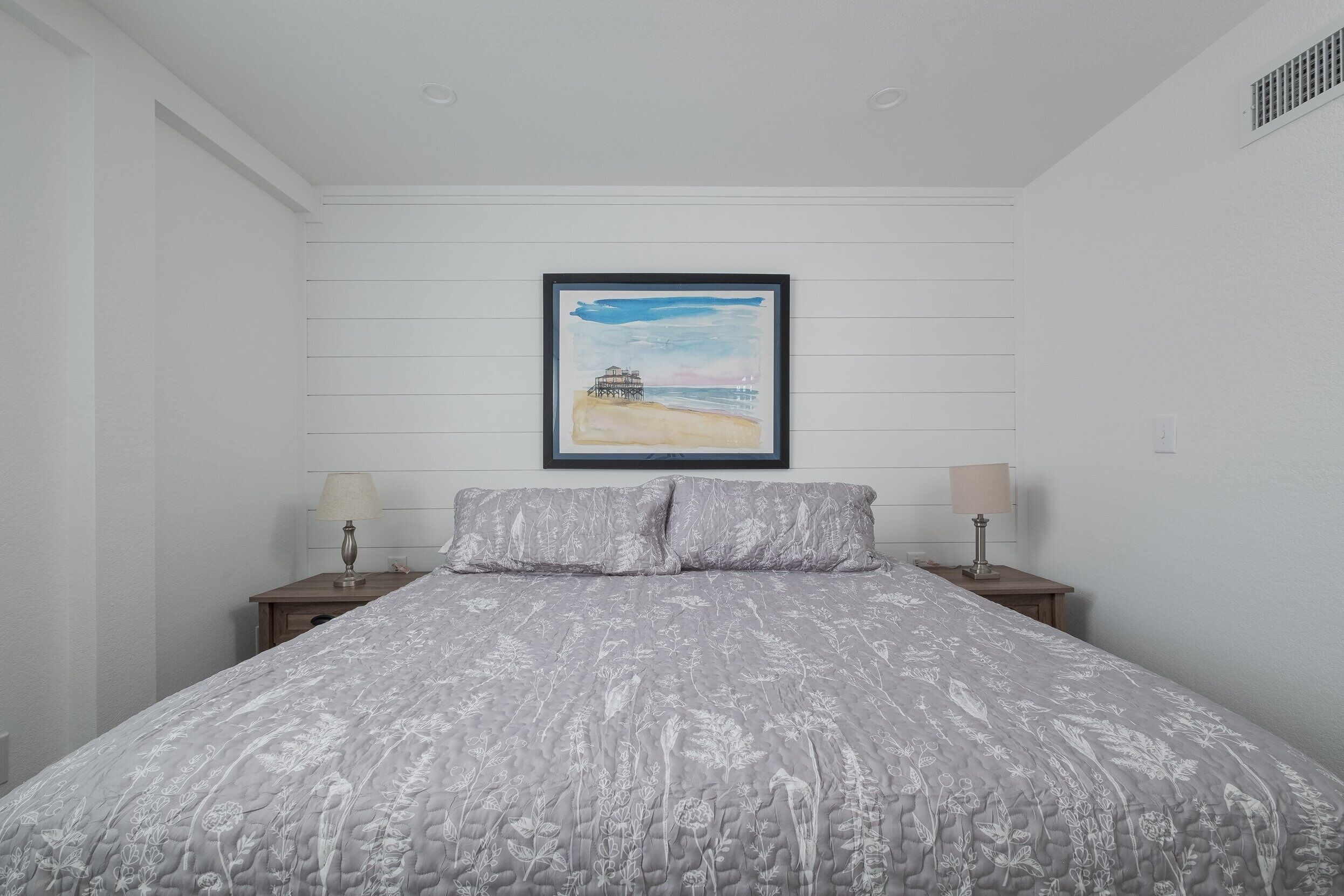 Une chambre avec un grand lit, deux tables de chevet avec des lampes et, au mur, un tableau représentant une scène de plage.