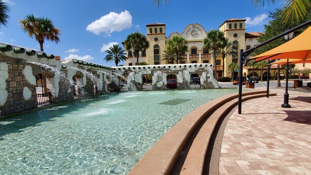 A clear pool with a stone wall and arches, surrounded by palm trees and a building with 'LA REINA' signage. - Hot Tub House Escape – 3BR in The Villages at Special Rates (The Villages)