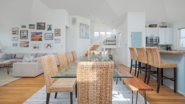 Ein geräumiges Wohnzimmer mit einem Glastisch, Korbstühlen und einer Couch. Es gibt eine Küchenzeile mit weißen Schränken und einer blauen Tür.
