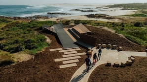 Un paseo marítimo con vista al océano, una playa arenosa y una zona verde con una estructura de madera.