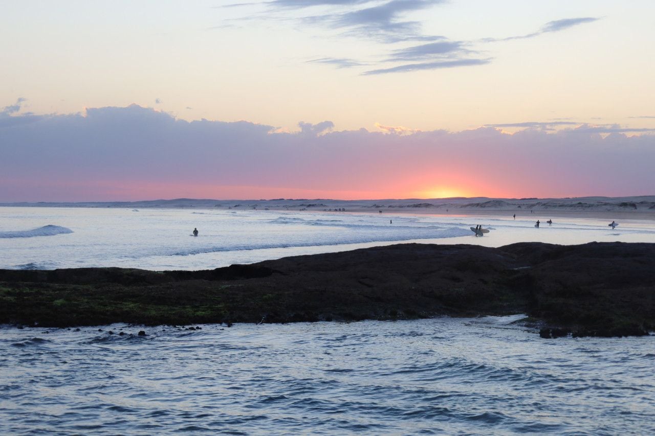 A beach scene with a sunset, rocky shoreline, and people in the water.