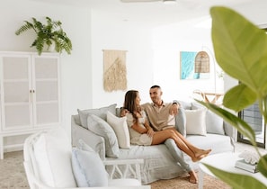Un couple est assis sur un canapé de couleur claire, dans une pièce lumineuse aux murs blancs, avec un meuble blanc et une plante suspendue.