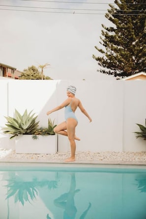 Une personne vêtue d’un maillot de bain bleu, debout près d’une piscine, avec un mur blanc et des plantes en arrière-plan.