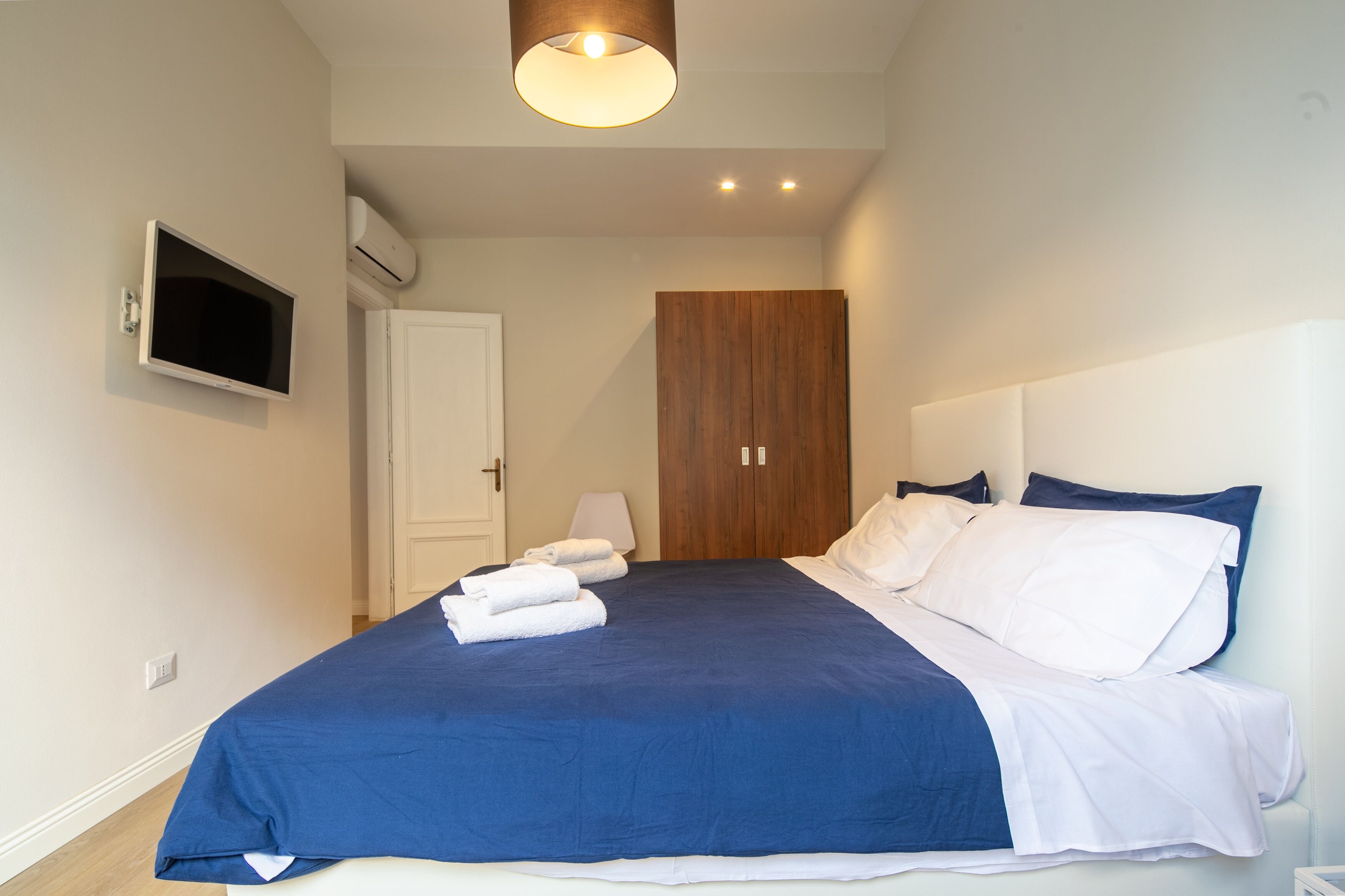 A bedroom with a bed, a television mounted on the wall, a wardrobe, and a door.