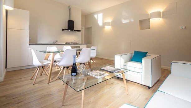 A modern living room with a glass coffee table, white chairs, a white sofa with a blue pillow, and a kitchen in the background. - Ponte Vecchio White by Mmega (Florence)