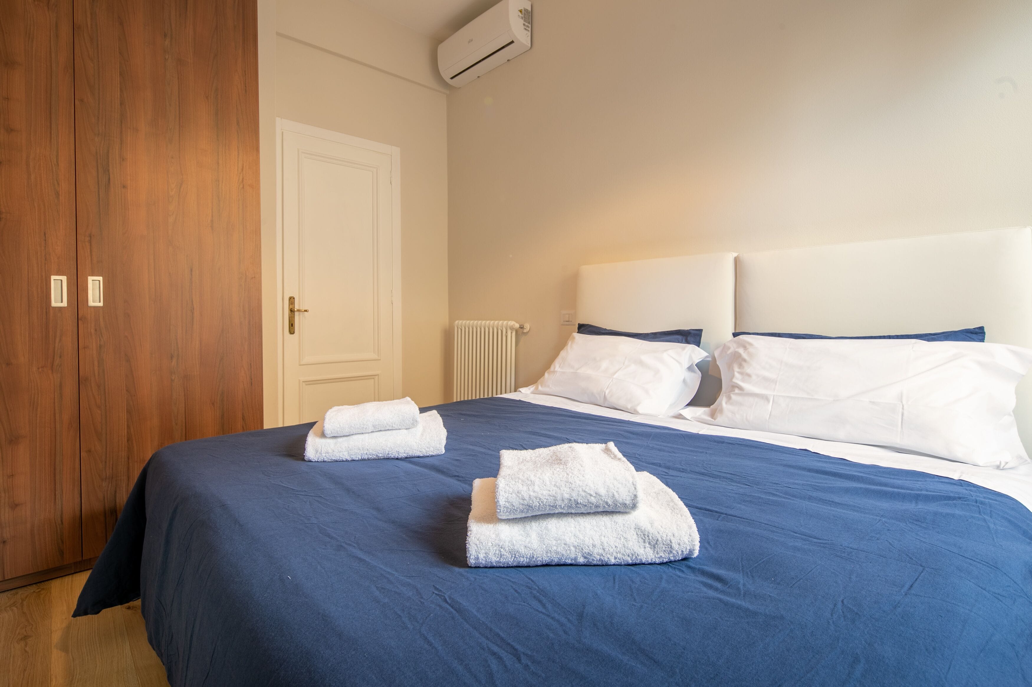 A neatly made bed with blue bedding and white pillows, a wooden wardrobe, and a white door.