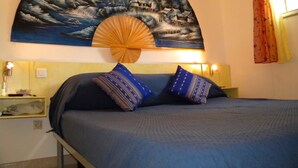 Een slaapkamer met een bed, twee kussens, een nachtkastje, een ventilator aan de muur en een schilderij boven het bed.