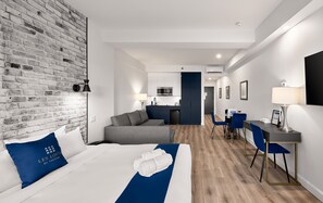 Ein Hotelzimmer mit Bett, Sofa, Schreibtisch, Stuhl, einer kleinen Küchenzeile und einer Ziegelwand.