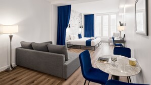 Et moderne hotelværelse med en grå sofa, en seng med blå gardiner, et skrivebord med stol og et rundt bord med lampe.