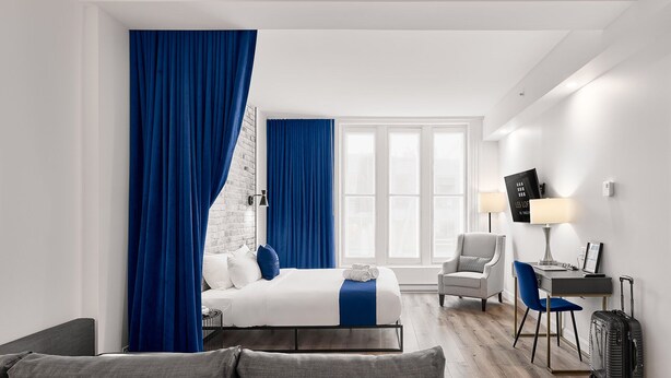 Et moderne hotelværelse med en seng, en grå sofa, et skrivebord med stol, et fjernsyn og en kuffert.
