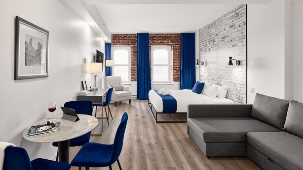 A modern hotel room with a bed, a grey sofa, a desk with a chair, a table with two chairs, and a brick wall. - Theater Lofts | Quebec, 606 (Québec City)
