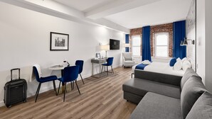 A modern hotel room with a bed, a desk with a chair, a small table with a chair, a suitcase, and a TV. - Theater Lofts | Quebec, 606 (Québec City)