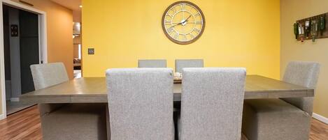 Un comedor con una mesa de madera, cuatro sillas tapizadas de gris y una pared amarilla con un reloj.