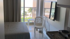 Une chambre d’hôtel comprenant un lit, une petite kitchenette, un coin repas avec une table et des chaises, et un balcon offrant une vue sur les palmiers et les immeubles.