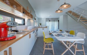 A modern kitchen with a red coffee machine, wooden shelves, and a dining area with a white table and yellow chairs. - Three Bedroom Melissi Villa ΙΙ with View Sea View (melissi)