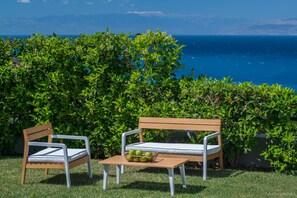 A wooden bench and a chair with cushions, placed on a grassy area with a view of the sea and a hedge in the background. - Three Bedroom Melissi Villa ΙΙ with View Sea View (melissi)