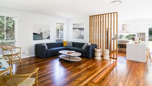 A modern living room with a dark blue sectional sofa, a coffee table, and a dining area with a kitchen in the background. - Blissful Bungalow - a luxurious escape (Culburra Beach)