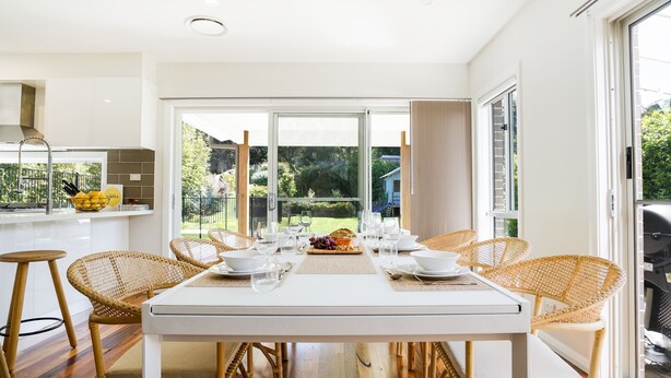 A modern dining area with a white table set for six, surrounded by wicker chairs, with a view of a garden through large windows. - Blissful Bungalow - a luxurious escape (Culburra Beach)