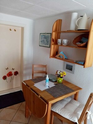 Un petit coin repas avec une table et des chaises en bois, une petite étagère avec des tasses et un cadre photo, et une porte décorée de motifs de coquelicots.