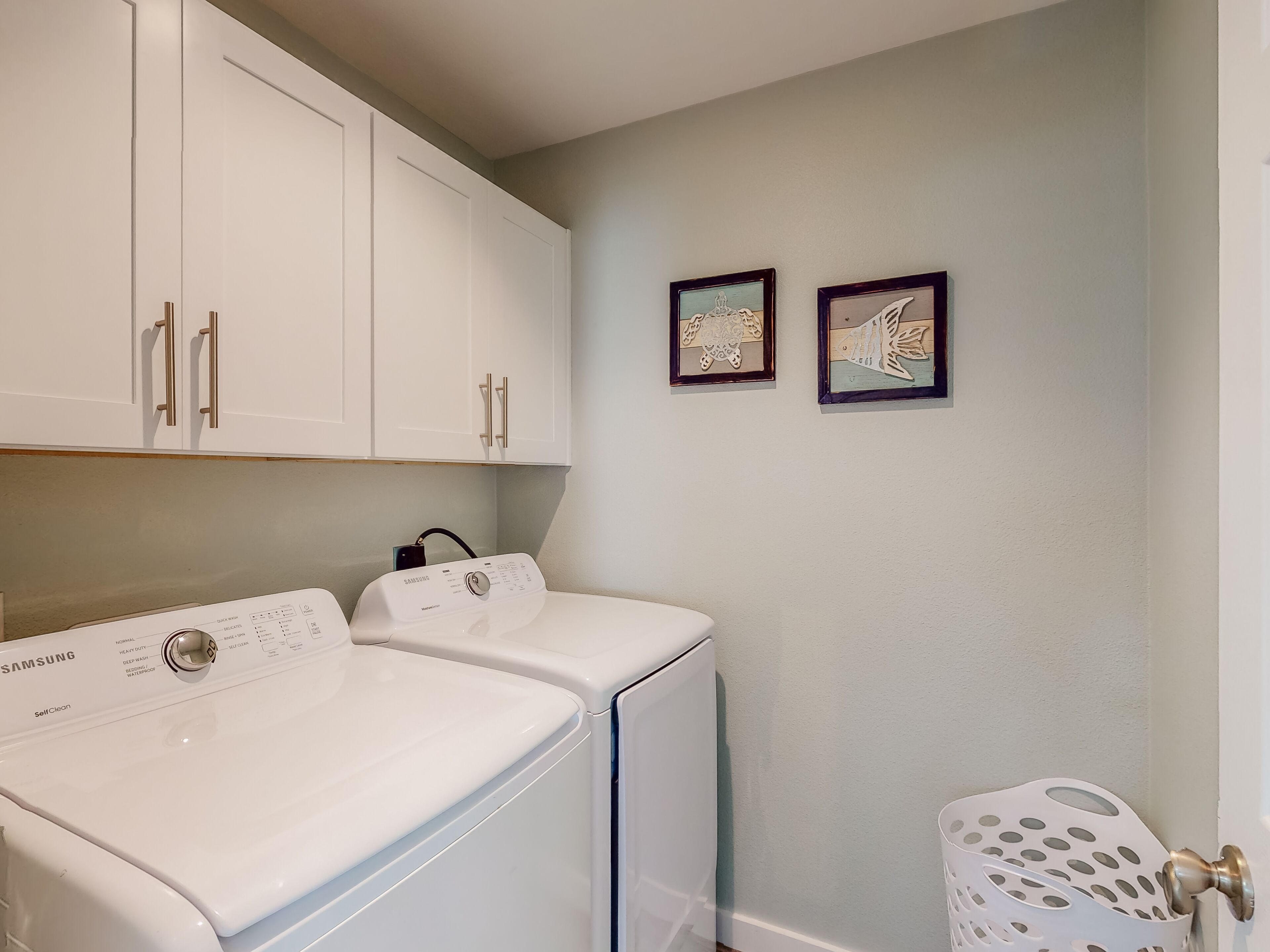 A laundry area with two white washers and dryers, a white laundry basket, and two framed pictures on the wall.