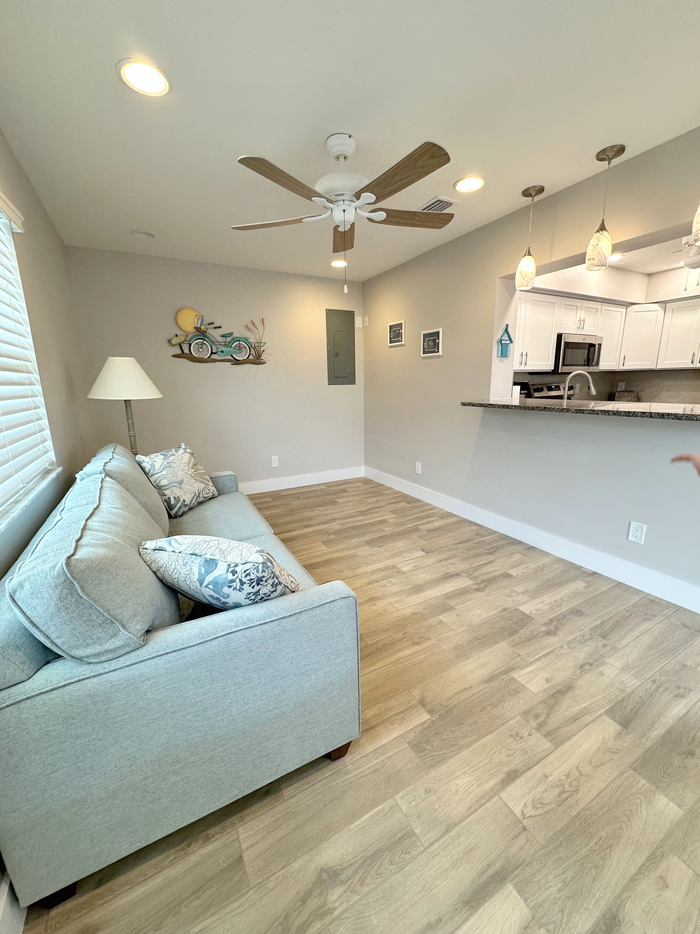 A modern living room with a light blue sofa, a ceiling fan, and a kitchen area with white cabinets and a microwave.