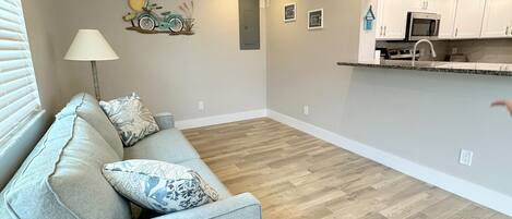 A modern living room with a light blue sofa, a ceiling fan, and a kitchen area with white cabinets and a microwave.
