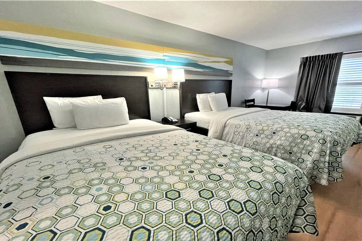 A hotel room with two beds, each with a patterned bedspread, a nightstand with a lamp, and a window with curtains.