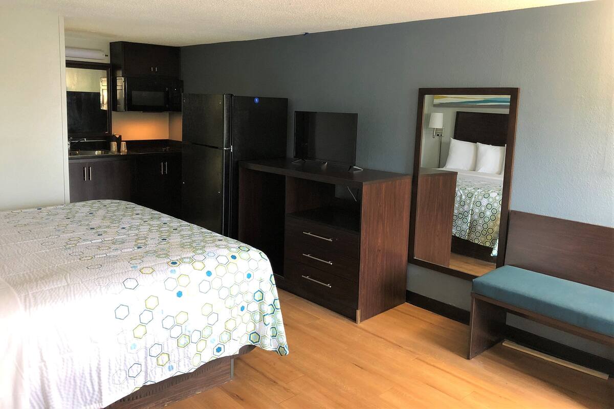 A hotel room with a bed, a desk with a computer, a mirror, a bench, and a kitchenette with a refrigerator and microwave.