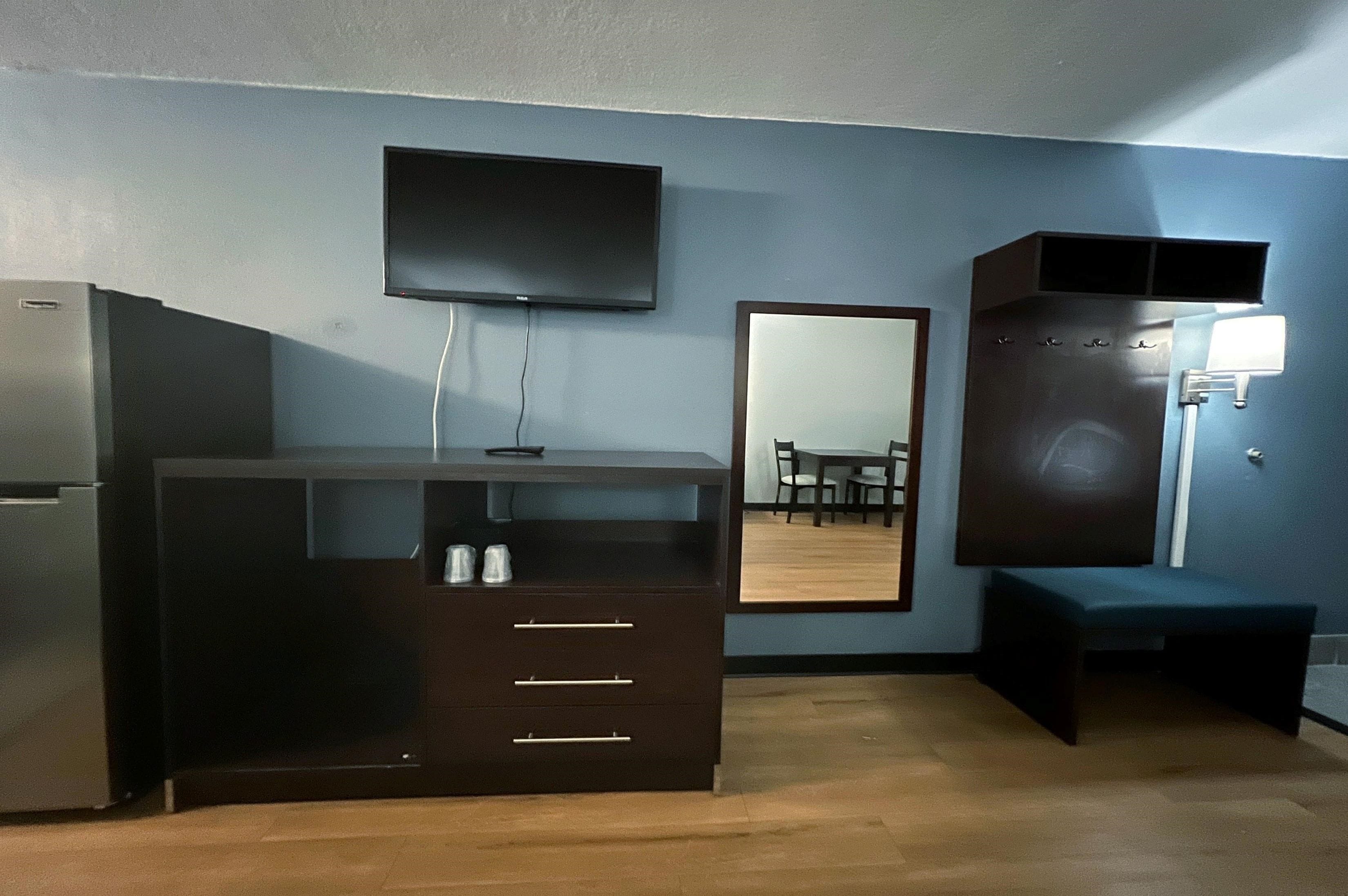 Una habitación compacta con kitchenette, un televisor de pantalla plana en la pared, un espejo grande que refleja un comedor y un banco.