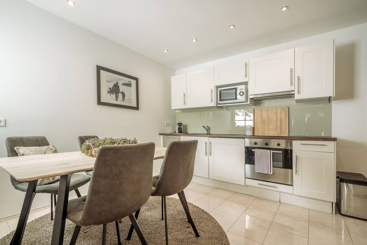 A modern kitchen with white cabinets, a microwave, and an oven. There is a dining table with two chairs and a rug.